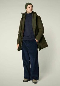 Man draagt een olijfgroene parka, een marineblauwe kabelgebreide trui, wijde marineblauwe broek, olijfgroene muts en beige schoenen, staand met één hand in de zak.