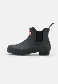 Bottines en caoutchouc noir avec panneaux élastiques sur les côtés, semelle texturée pour une meilleure adhérence et une languette pour un enfilage facile. Détail du logo rouge sur le dessus.