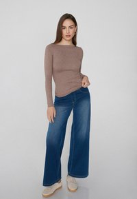 Top marrón de manga larga con escote barco, combinado con jeans de pierna ancha de color azul y zapatillas blancas con suela de color claro.