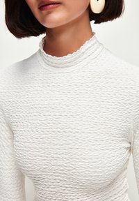 Top bianco texturizzato con collo alto e scallopato. Fit aderente con un motivo ondulato. Orecchini grandi e di tendenza dalla forma curva.