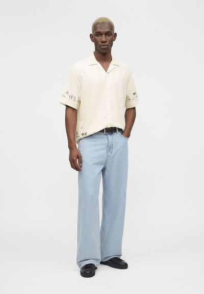 Άνδρας με κοντά ξανθά μαλλιά που φοράει κρεμ πουκάμισο με κοντά μανίκια, κουμπωτό, με μαύρα τροπικά σχέδια και ανοιχτό μπλε παντελόνι με φαρδιές μπατζάκια.