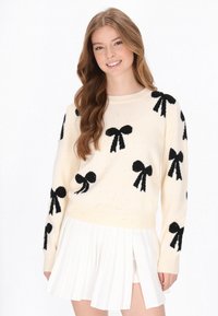 Krämfärgad tröja med svarta rosettmönster och subtil glans, rund hals, och långa ärmar, i kombination med en vit plisserad kjol.
