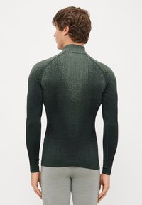 Longsleeve'owa koszulka sportowa w ciemnozielonym kolorze, z teksturowanej tkaniny o gradientowym wzorze. Wysoka kołnierz i dopasowany krój zwiększają elastyczność.