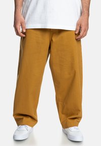 Gule bomuldsbukser med bredde ben, der har en glat tekstur og en afslappet pasform, parret med hvide sneakers. Enkel design uden synlige mønstre.