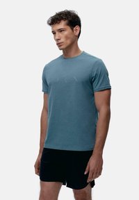 Kortermet t-skjorte med rund hals i dempet teal-stoff. Glatt tekstur med subtile mønstre. Kombinert med svarte shorts.