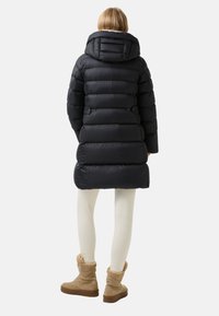 Černý puffer kabát se silným, prošívaným designem, s vysokým límcem a střední délkou. Nosit s bílými legínami a béžovými botami.