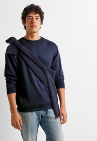 Sudadera azul marino con un diseño texturizado en los hombros, mangas largas y cuello redondo, combinada con unos vaqueros azul claro.
