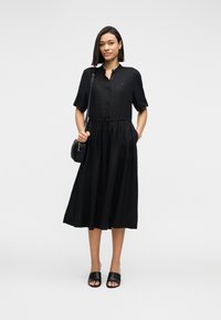 Γυναίκα που φοράει μαύρο μίντι φόρεμα με κουμπιά και κοντά μανίκια, μαύρες σανδάλες σλάιντ και μικρή μαύρη τσάντα ώμου, στέκεται μπροστά σε λευκό φόντο.