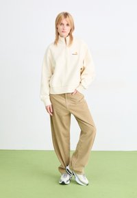 Felpa crema con zip e logo ricamato, abbinata a pantaloni in corduroy beige larghi e sneaker grigie. Design semplice, vestibilità rilassata.