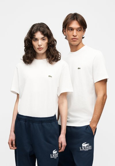 Два модела, облечени в бели тениски Lacoste и тъмносини спортни панталони Lacoste Paris, стоят един до друг на еднообразен фон.