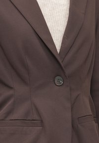 Brūns bleizera ar izgrieztu atloku, vienu melnu pogu un divām priekšējām kabatām. Gluda auduma ar smalkām šuvēm un minimālu tekstūru.