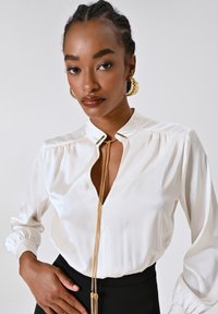 Blusa in satin bianco con colletto alto, scollatura a V profonda e dettaglio in catena dorata, abbinata a pantaloni neri tailored. Texture liscia.