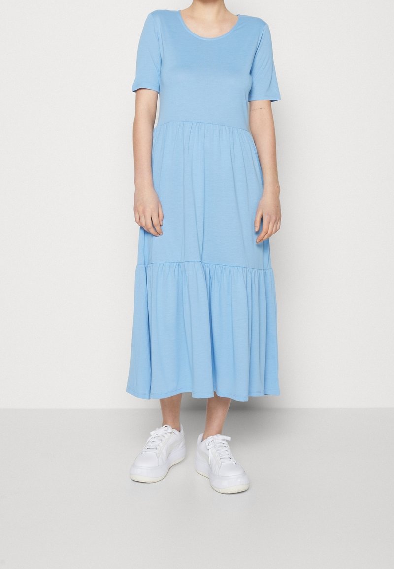 Femme portant une robe midi bleu clair à manches courtes avec une jupe à volants et des baskets blanches à lacets, debout devant un fond uni.
