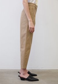 Béžové kalhoty s vysokým pasem a úzkým střihem, se rovnými nohavicemi, bočními kapsami a jemnými švy, doplněné černými muly.