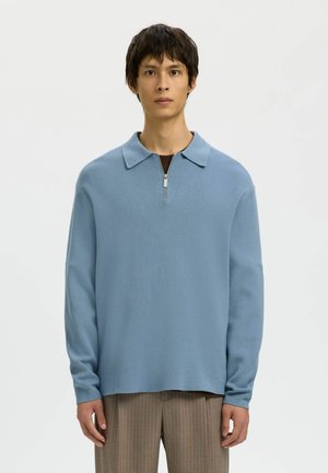 Jeune homme debout portant un pull bleu clair à col zippé et un pantalon marron à fines rayures sur fond blanc uni.