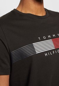 Man wearing black t-shirt featuring horizontal striped design in navy, white, and red with "Tommy Hilfiger" text on chest.