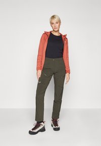 Moleton laranja com fecho, camisa preta, calças cargo verde-oliva com bolsos laterais e botas de caminhada cinzentas com detalhes vermelhos.