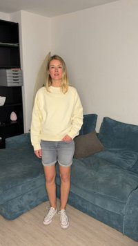 Γυναίκα με ξανθά μαλλιά που φοράει κρεμ φούτερ, γκρι σορτς και λευκά αθλητικά, στέκεται σε ένα σαλόνι κοντά σε έναν μπλε καναπέ με μαξιλάρια.