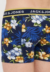 Jūras spēku bokseršorti ar ziedu rakstu, kas satur zilos un dzeltenos hibiskus, zaļas lapas un kontrastējošu jostasvietu ar logo.