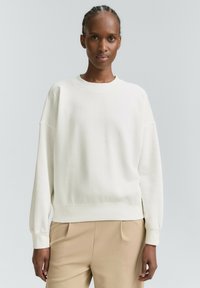 Ženska z opletenimi lasmi, oblečena v preprosto belo srajco z dolgimi rokavi in bež hlače, na svetli nevtrali podlagi.