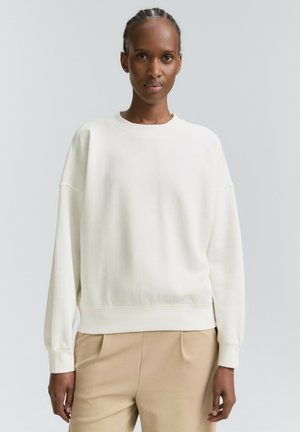 Vrouw met een gevlochten haar, gekleed in een eenvoudige witte sweater met lange mouwen en beige broek, tegen een lichte neutrale achtergrond.