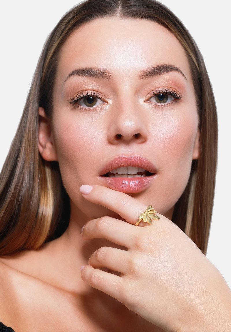 Gouden ring met een bladontwerp op de vinger, zachte textuur, glanzende afwerking. Model toont de ring terwijl hij/zij zachtjes de kin op de hand steunt. Neutrale achtergrond.