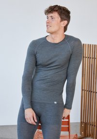 Harmaa pitkähiha thermo-paita, joka on valmistettu pehmeästä, joustavasta kankaasta. Siinä on pyöreä pääntie ja resorit hihansuissa. Tiimalasimallinen leikkaus, jota korostaa ompelu.