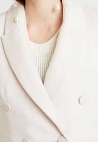 Blazer doble faz de color beige claro en tejido texturizado, con grandes botones redondos y cuello con muesca, superpuesto a un suéter crema.