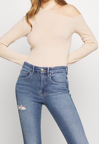 Vaalean beige ribattu bodysuit, jossa on olkapäiltä paljastava malli, yhdistettyinä sinisiin korkeavyötäröisiin farkkuihin, joissa on revennyt yksityiskohta polvessa.