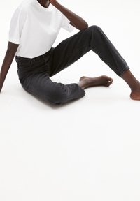 Personne assise par terre portant un t-shirt blanc et un jean noir, avec une jambe pliée et un bras reposant sur le genou plié.
