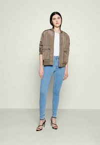 Mulher de pé, vestindo uma jaqueta bomber taupe, uma camisa branca, jeans justos azul-claro e sandálias de salto alto pretas, contra um fundo simples.