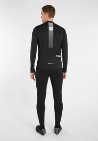 Zwarte lange mouw fietsjersey met reflecterende strepen, een achterzak en een aansluitend ontwerp. Gekleed met zwarte legging en fietsschoenen.