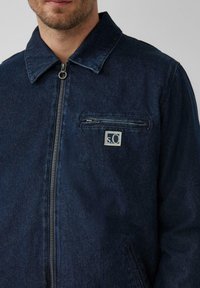 Dunkelblaue Jeansjacke mit Reißverschluss, spitzem Kragen und einer Brusttasche mit rechteckigem Logo-Patch. Angenehme Textur.