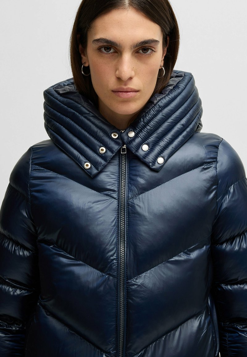 Kurtka puchowa w ciemnym odcieniu granatu z wzorem w zygzak, z wysokim, pikowanym kołnierzem i srebrnymi guzikami jako dodatkiem. Gładka, błyszcząca tekstura.