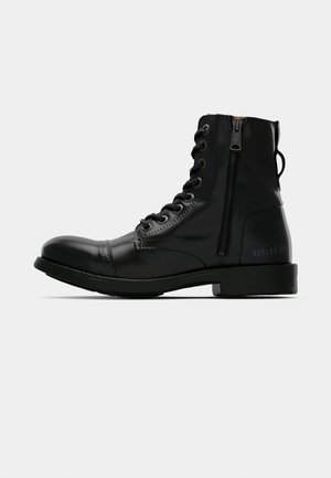 Svart skinn ankel støvel med rund tå, snøring foran, glidelås på siden og flat såle. Har preget merkevare på hælen.