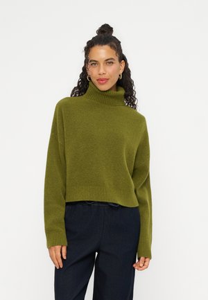 Femme aux cheveux bouclés attachés en chignon, portant des boucles d’oreilles anneaux dorés, un pull à col roulé vert olive et un pantalon bleu foncé, debout devant un fond blanc.