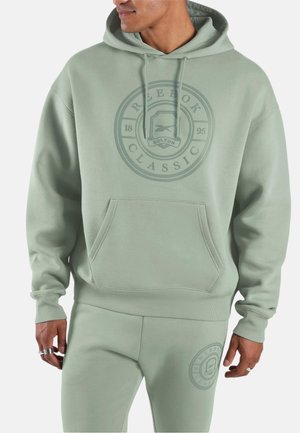 Man draagt lichtgroene Reebok Classic hoodie en bijpassende sweatpants met logo's, handen ontspannen langs het lichaam.