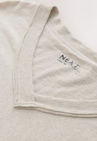 T-shirt beige doux à col en V avec coutures visibles et étiquette de marque "NEXT" à l'intérieur du col, taille V.MED CT XS.
