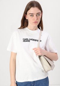 Biela tričko s čiernym nápisom "KARL LAGERFELD JEANS", spárované s modrými džínsami a svetlošedou kabelkou držiacou v jednej ruke.