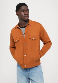 Chaqueta color óxido hecha de un tejido texturizado, con cuello, dos bolsillos en el pecho, cierre de botones y puños de canalé, combinada con jeans claros.