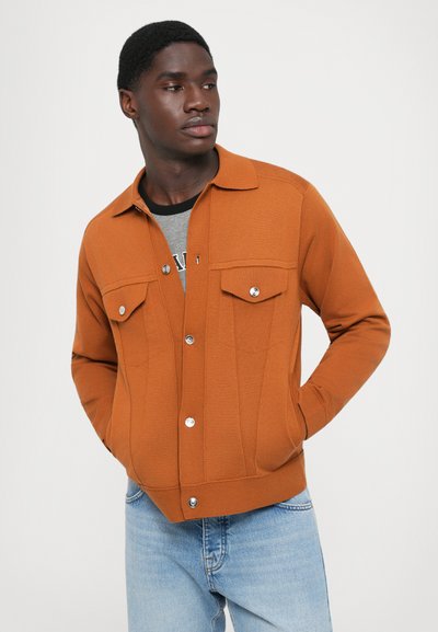 Brun cardigan jakke med krave, to brystlommer, sølvknapper, ribbet tekstur og en tætsiddende pasform, parret med lyseblå jeans.
