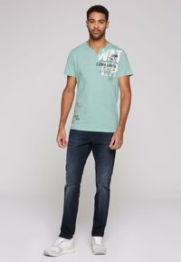 Camiseta de algodón verde claro con escote en forma de V, con gráficos y texto impresos. Combinada con jeans azul oscuro y zapatillas blancas.
