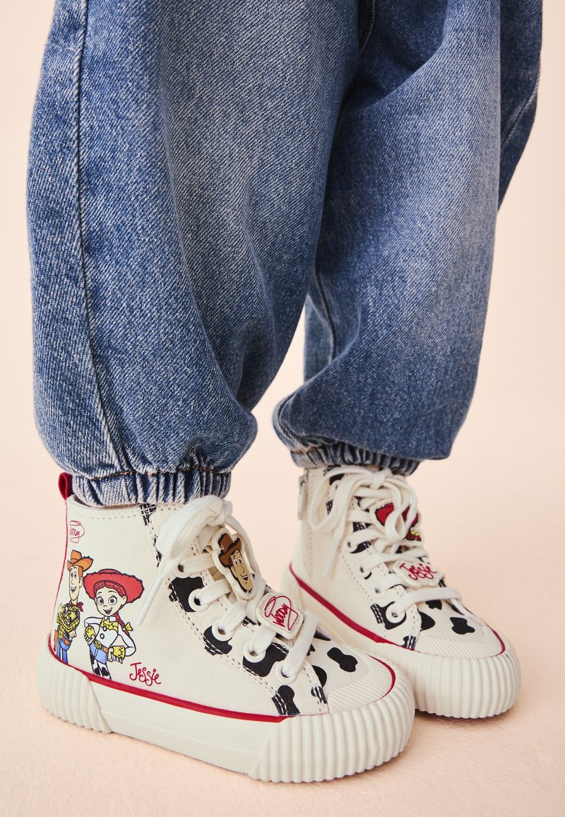 Gros plan d’un enfant portant un pantalon en jean bleu et des baskets montantes blanches avec les personnages Woody et Jessie de Toy Story, ainsi que des taches noires de vache.