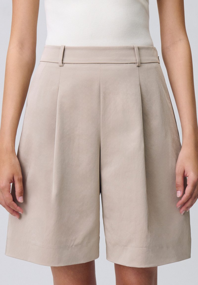 Shorts beige chiari a vita alta realizzati in tessuto morbido. Presentano delle pieghe sul davanti e una vestibilità dritta, che termina appena sopra il ginocchio.