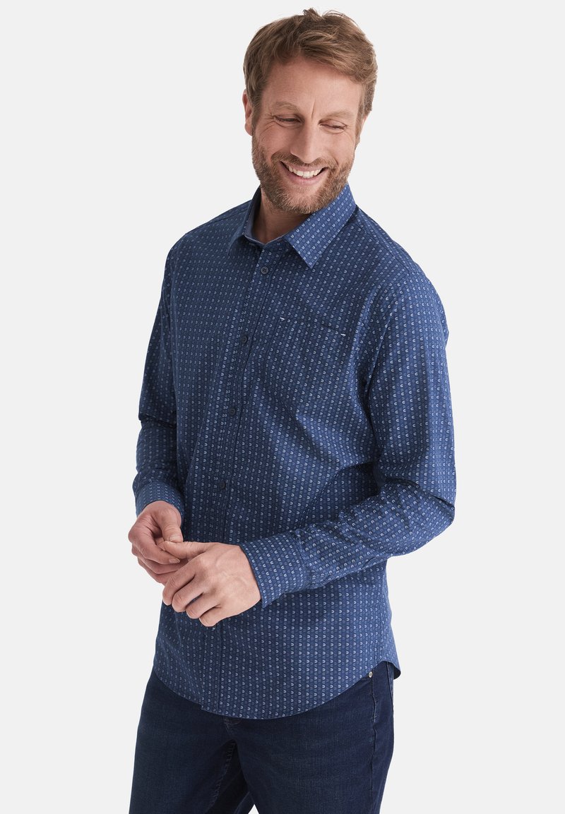 Dunkelblaues Hemd mit Knopfleiste, kleinem Muster, langen Ärmel und einer Brusttasche, aus leichtem Stoff. Kombiniert mit dunkelblauen Jeans.