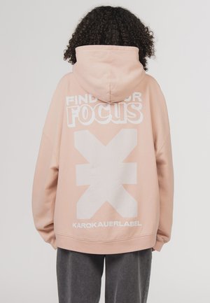 Person with curly hair wearing a light pink hoodie with white text and a large graphic on the back and dark gray pants.