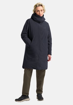 Marineblauwe lange parka met een hoge kraag, gemaakt van een gladde, waterafstotende stof. Twee zijzakken en een losse pasvorm. Draagt met olijfgroene broek.