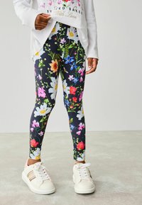 Blommiga leggings med svart bakgrund och flerfärgade blommor, tillverkade av elastisk tyg. Kombineras med vita sneakers med rosa detaljer.