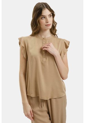 Młoda kobieta w jasno-brązowej bluzce bez rękawów z falbankami na ramionach i dopasowanych luźnych spodniach, patrząca na bok z ręką przy kołnierzu.