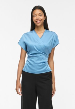 Femme debout et souriante portant un chemisier cache-cœur bleu clair à manches courtes et un pantalon noir à jambes larges sur fond blanc.